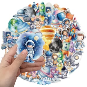 May include: A collection of watercolour-style stickers featuring space themes, including astronauts, rockets, planets, moons, and stars in various colours. One sticker showing an astronaut in a blue starry sky is held in the foreground.