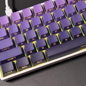 May include: A close-up of a mechanical keyboard with purple keycaps and a white frame. The keys are arranged in a standard QWERTY layout, with yellow backlighting visible between the keys. The keyboard is connected to a white cable.