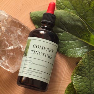 May include: A dark amber glass bottle with a red dropper top labelled "Comfrey Tincture" sits on a wooden surface. The label includes the product name and other text. Large green leaves and a clear quartz crystal are also in the image.
