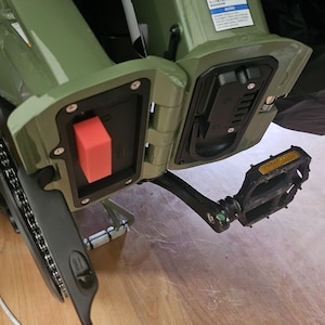 May include: Close-up of a green electric bicycle frame with black pedals and a chain. The frame has a red rectangular button and a black compartment. The pedals have yellow reflectors.