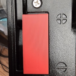 May include: A black textured metal latch mechanism featuring a rectangular red plastic insert. The assembly is secured with two visible Phillips head screws and includes embossed circular icons representing a four-quadrant grid and a horizontal line.
