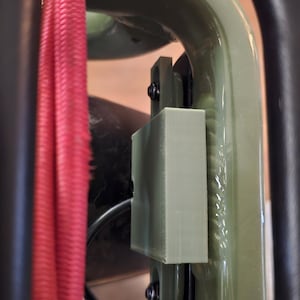 May include: Close-up of a section of a bicycle frame with a red cable and a light green rectangular component attached with black screws. The frame is olive green and black.
