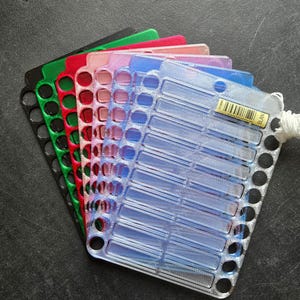 Puede incluir: Un conjunto de bobinas rectangulares de plástico para hilo de bordar apiladas en negro, verde, rojo, rosa, rosa claro, morado y purpurina transparente. Cada bobina presenta ranuras horizontales y orificios circulares laterales con etiqueta.