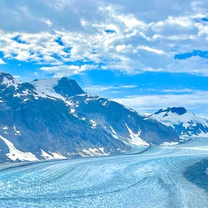 May include: A scenic landscape featuring a glacier winding through a valley, flanked by snow-capped mountains under a bright blue sky dotted with fluffy white clouds. The glacier's surface is a striking blue.