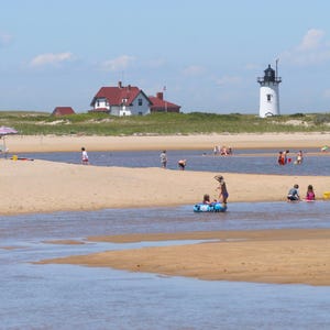 Puede incluir: Una escena de playa soleada con un faro blanco y una casa con techo rojo. La gente disfruta del agua y la arena. El cielo es azul con algunas nubes. El agua es una mezcla de azul y marrón.