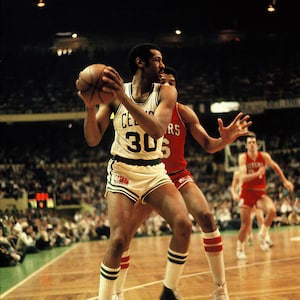 May include: A vintage action shot of a basketball game. A player in a white and green Celtics uniform, number 30, dribbles the ball. Another player in a red uniform defends. The background shows a packed stadium and championship banners.
