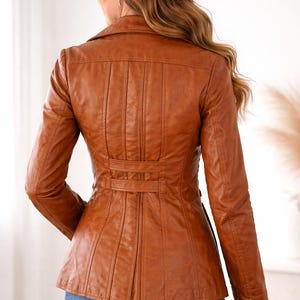 May include: A brown leather jacket with a tailored fit. The jacket features a notched collar, vertical seams on the back, and a belt detail at the waist. The sleeves are long, and the jacket is worn with blue jeans.
