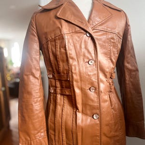May include: A vintage brown leather jacket with a collared neckline and button-up closure. The jacket features decorative pleats and small buckle details. The jacket is displayed on a mannequin.