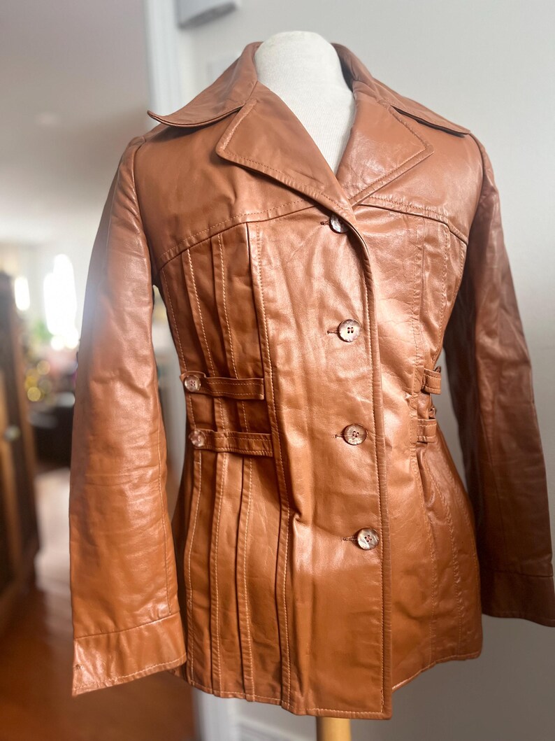 May include: A vintage brown leather jacket with a collared neckline and button-up closure. The jacket features decorative pleats and small buckle details. The jacket is displayed on a mannequin.