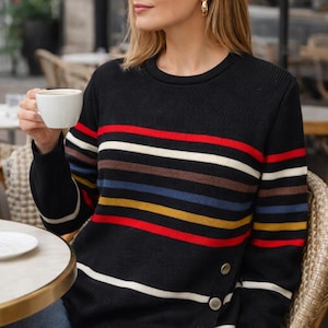 Peut inclure: Pull noir à rayures horizontales rouges, blanches, marron, bleues et jaunes. Le pull a un col rond et des boutons décoratifs sur le côté. La personne est assise à une table de café.