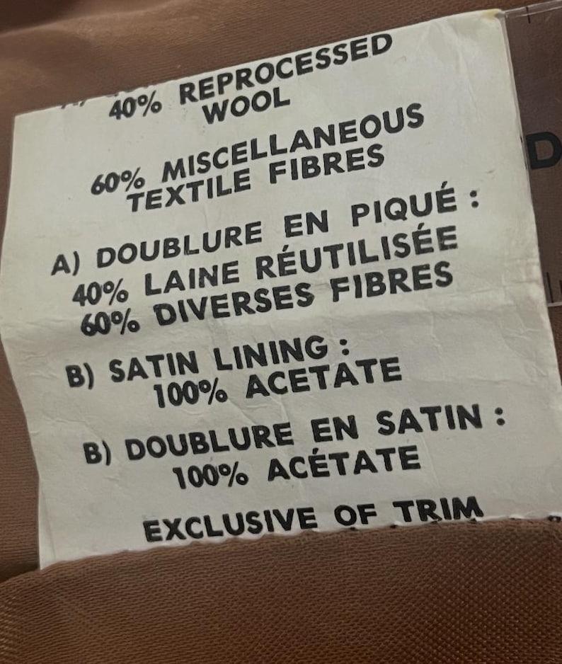 May include: A close-up of a tag with text detailing the fabric composition of a garment. The tag lists percentages of wool, textile fibers, and acetate, along with French terms for lining and trim. The tag is against a brown fabric background.