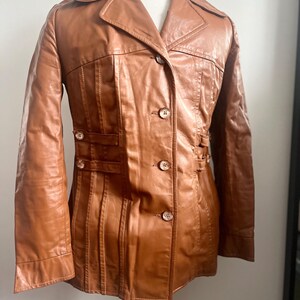 May include: A brown leather jacket with a notched collar, button closures, and decorative stitching. The jacket features a tailored fit with vertical seams and small button details on the waist. The jacket is displayed on a mannequin.