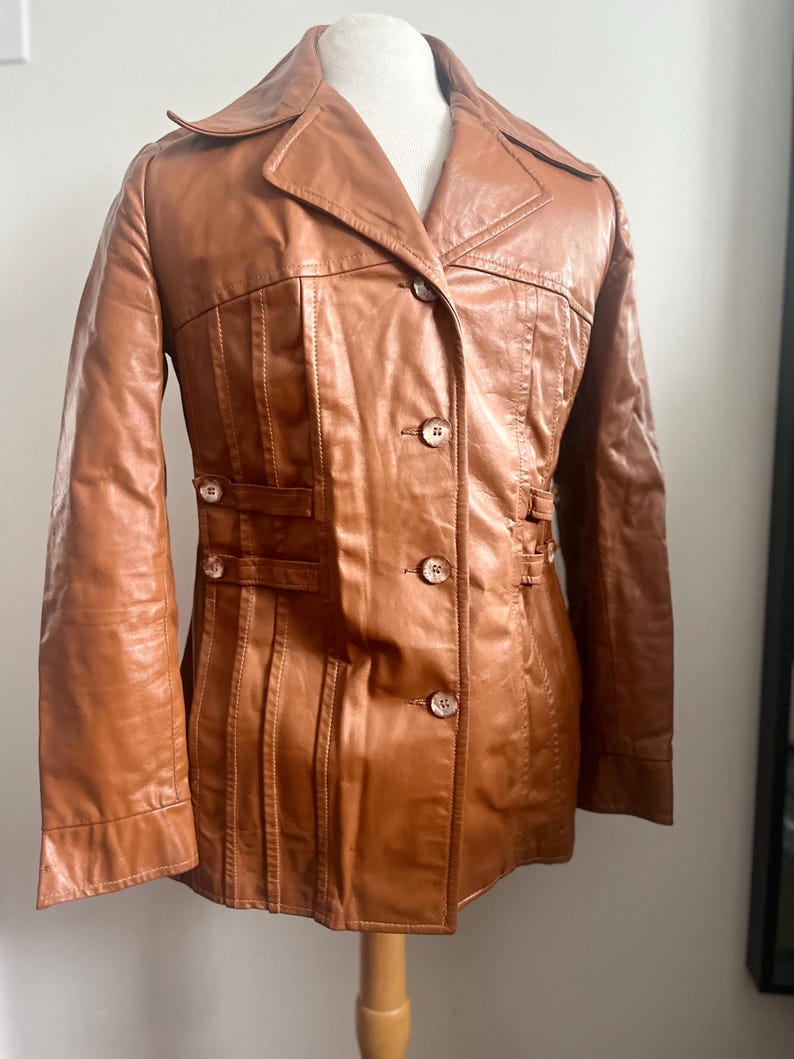 May include: A brown leather jacket with a notched collar, button closures, and decorative stitching. The jacket features a tailored fit with vertical seams and small button details on the waist. The jacket is displayed on a mannequin.