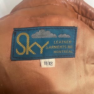 May include: Close-up of a brown leather jacket interior with a blue label that reads "Sky Leather Garments Inc Montreal". The label also has the size "11/12". The jacket's lining is a light brown color.