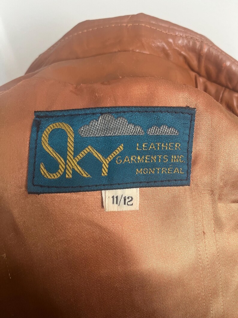 May include: Close-up of a brown leather jacket interior with a blue label that reads "Sky Leather Garments Inc Montreal". The label also has the size "11/12". The jacket's lining is a light brown color.