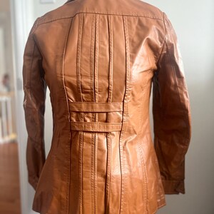 May include: A tan leather jacket with a classic design. The jacket features a collared neckline, long sleeves, and a fitted waist. The back of the jacket has vertical pleats and a decorative belt detail. The jacket is displayed on a wooden mannequin.