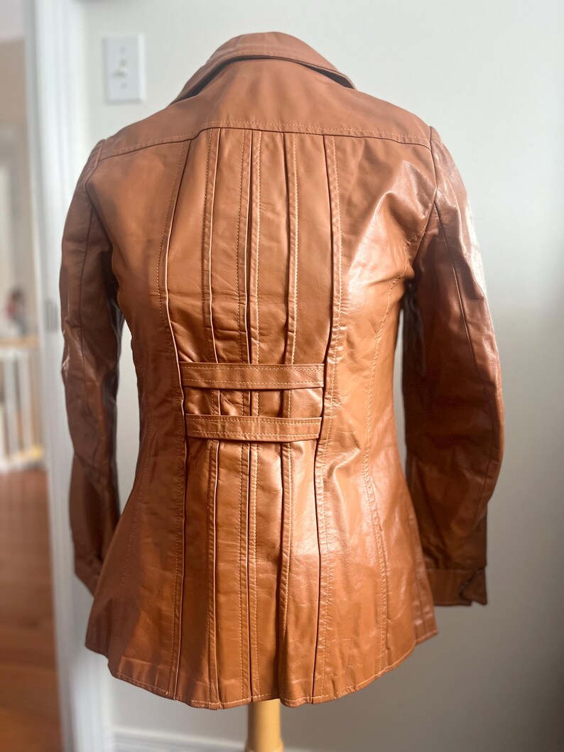 May include: A tan leather jacket with a classic design. The jacket features a collared neckline, long sleeves, and a fitted waist. The back of the jacket has vertical pleats and a decorative belt detail. The jacket is displayed on a wooden mannequin.