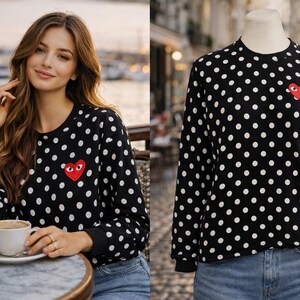 May include: Black long-sleeved sweatshirt with white polka dots and a red heart-shaped patch. The sweatshirt is worn by a person and displayed on a mannequin. The person is sitting at a table with a cup of coffee.