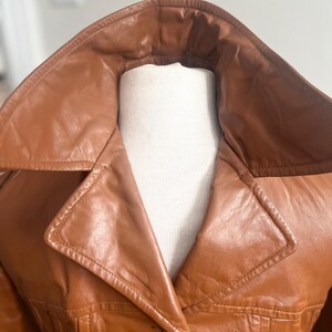 May include: A close-up of a brown leather jacket with a wide collar. The jacket is displayed on a white mannequin. The leather appears to be smooth and has a slight sheen. The stitching details are visible along the seams and collar.