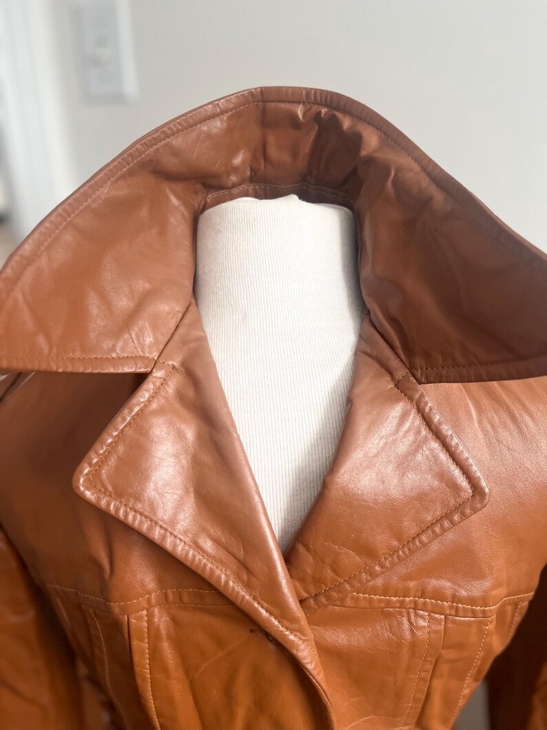 May include: A close-up of a brown leather jacket with a wide collar. The jacket is displayed on a white mannequin. The leather appears to be smooth and has a slight sheen. The stitching details are visible along the seams and collar.