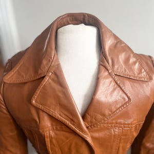 May include: A vintage brown leather jacket with a wide collar and button closure. The jacket has a tailored fit and visible stitching details. The leather appears to have a slight sheen, suggesting it's well-maintained. The jacket is displayed on a mannequin.