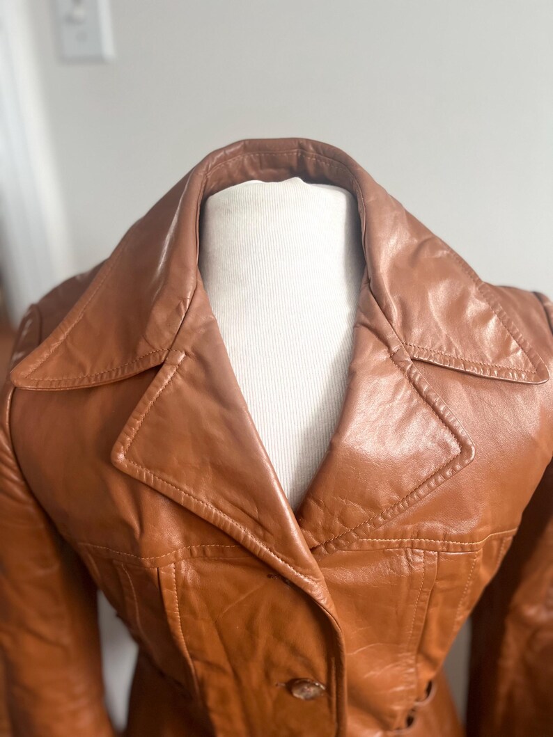May include: A vintage brown leather jacket with a wide collar and button closure. The jacket has a tailored fit and visible stitching details. The leather appears to have a slight sheen, suggesting it's well-maintained. The jacket is displayed on a mannequin.
