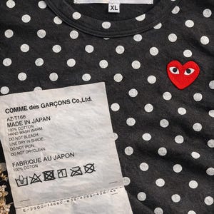 May include: Dark grey cotton t-shirt with white polka dots and a red heart logo with eyes. The shirt has a crew neck and the text "PLAY COMME des GARCONS" on the label. The size is XL. A tag with washing instructions is also visible.