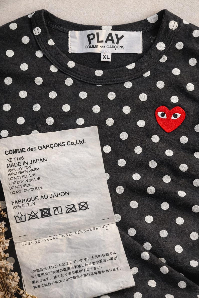 May include: Dark grey cotton t-shirt with white polka dots and a red heart logo with eyes. The shirt has a crew neck and the text "PLAY COMME des GARCONS" on the label. The size is XL. A tag with washing instructions is also visible.