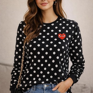May include: Black long-sleeved top with white polka dots and a red heart motif. The top is worn with blue denim jeans. A black handbag with a gold chain is visible.