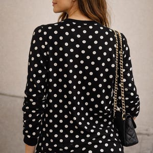 May include: A black top with a white polka dot design. A small black quilted handbag with a gold chain strap is worn over the shoulder. The person is wearing blue jeans.
