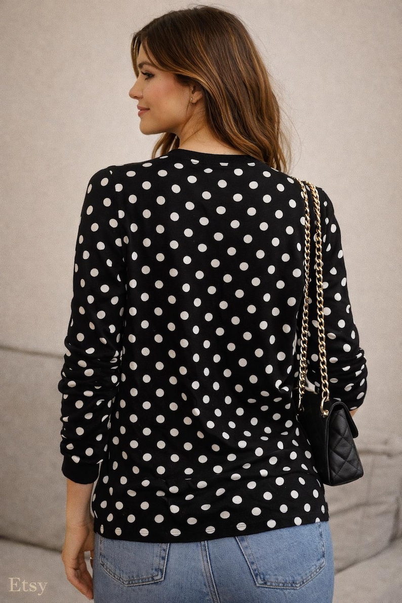 May include: A black top with a white polka dot design. A small black quilted handbag with a gold chain strap is worn over the shoulder. The person is wearing blue jeans.