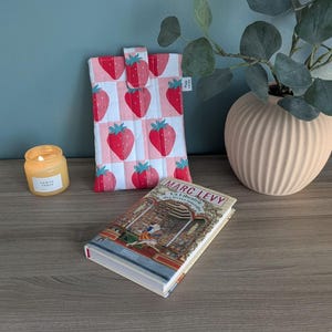 Può includere: Una custodia per libri trapuntata con un motivo a fragole, un libro con il titolo "Marc Levy", una candela accesa in un barattolo di vetro e un vaso decorativo con vegetazione.