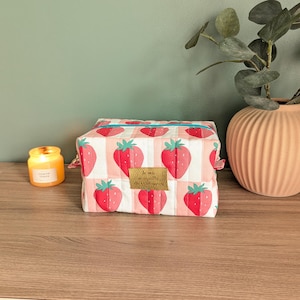 Peut inclure: Une trousse de maquillage carrée matelassée avec un motif de fraises. Le sac présente des fraises rouges sur un fond à rayures blanches et roses. Une bougie allumée et un vase pêche avec de la verdure sont en arrière-plan.