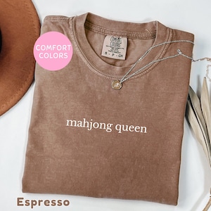 May include: Espresso-colored t-shirt with the text "mahjong queen" in white. The shirt has a crew neck and short sleeves. A silver necklace with a pendant is draped across the shirt. The shirt is labeled "Comfort Colors".