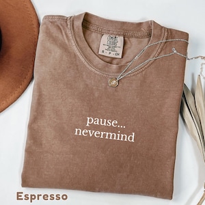 May include: An espresso-colored short-sleeve t-shirt with a crew neckline. The shirt features the white text "pause... nevermind" printed on the chest. A Comfort Colors brand label is visible at the inner neck, indicating size S, and a silver necklace.