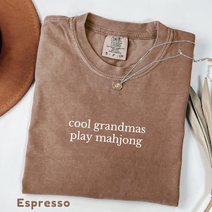 May include: A brown short-sleeved t-shirt with a crew neckline and the white text "cool grandmas play mahjong" printed on the chest. The shirt features a Comfort Colors brand label at the inner neck and is paired with a silver-toned pendant necklace.