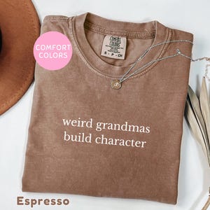 May include: A brown short-sleeved t-shirt with the text "weird grandmas build character" printed in white on the chest. The shirt features a Comfort Colors brand label at the inner neckline and is paired with a thin silver chain necklace.