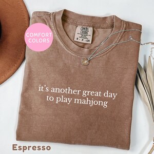 May include: Espresso-colored t-shirt with the text "it's another great day to play mahjong" in white. The shirt has a crew neck and short sleeves. A silver necklace with a pendant is draped across the shirt.