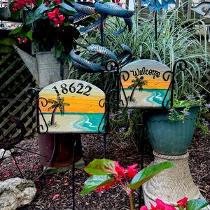 May include: Two decorative garden signs with a beach scene painted on them. One sign displays the number "18622" and the other says "Welcome". The signs are mounted on black metal stands and surrounded by plants and flowers.