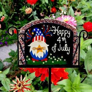 May include: A black garden stake with the text "Happy 4th of July" and a gnome wearing a patriotic hat. The gnome holds a star with "USA" on it. The stake is surrounded by red, orange, and pink flowers.