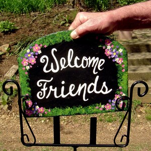 May include: A black slate welcome sign with white lettering that reads "Welcome Friends". The sign is decorated with painted pink, purple, and white flowers and green foliage. The sign is mounted on a black metal stand.