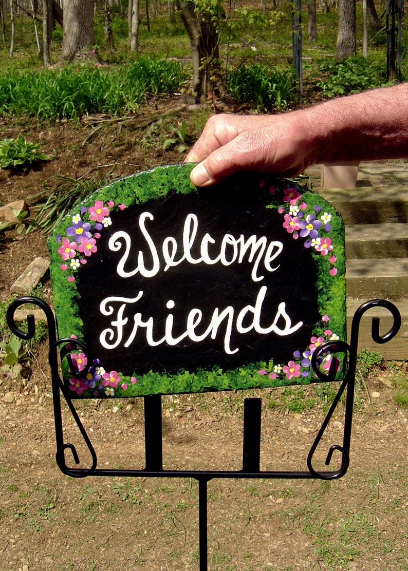 May include: A black slate welcome sign with white lettering that reads "Welcome Friends". The sign is decorated with painted pink, purple, and white flowers and green foliage. The sign is mounted on a black metal stand.