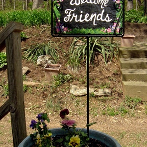 May include: A black metal sign with a green and brown background and white painted flowers and the words "Welcome Friends". The sign is attached to a black metal stand and is placed in a grey planter with green plants and purple and pink flowers.