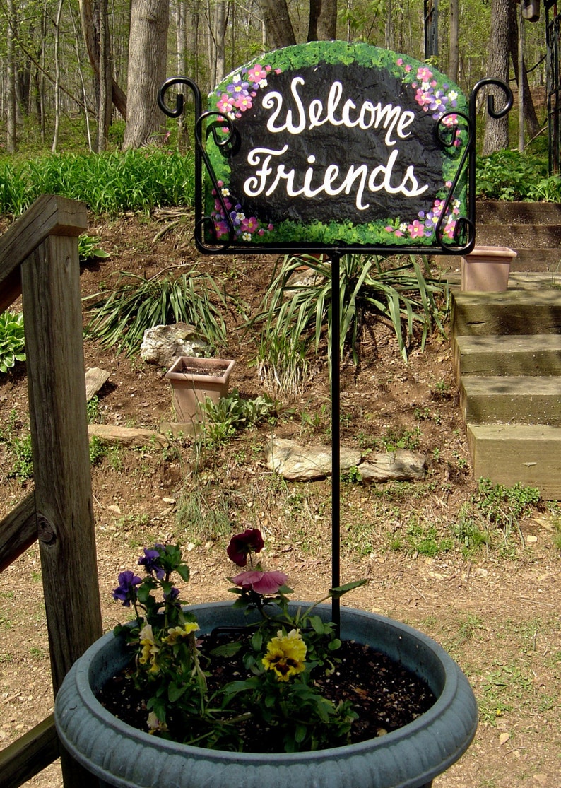 May include: A black metal sign with a green and brown background and white painted flowers and the words "Welcome Friends". The sign is attached to a black metal stand and is placed in a grey planter with green plants and purple and pink flowers.