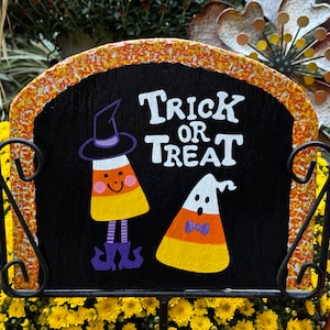 May include: Black Halloween sign with orange and yellow trim. The sign says "Trick or Treat" and features two candy corn characters, one wearing a witch's hat and the other a ghost.