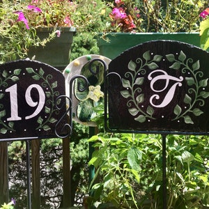 May include: Two black slate signs with white painted numbers and a letter, surrounded by green painted leaves, on black metal stands. The sign on the left says "19" and the sign on the right says "T".