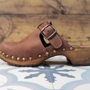 Può includere: Zoccolo in pelle marrone con suola in legno. La scarpa presenta una chiusura con fibbia ed è decorata con borchie color oro attorno alla suola. Lo zoccolo è posto su uno sfondo di legno.