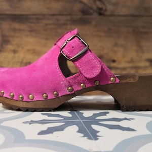 May include: A bright pink suede clog with a wooden sole. The shoe features a buckle closure and is adorned with brass-colored studs along the edge. The heel is approximately 2 inches high.