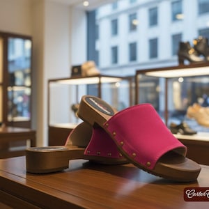 May include: A pair of pink clogs with wooden soles and a wide strap. The clogs are displayed on a wooden table in a shoe store. Other shoes are visible in the background, including boots and sneakers. The clogs have a heel height of approximately 2 inches.