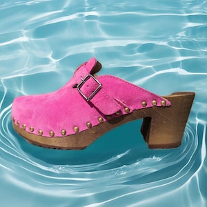 May include: A pink suede clog with a wooden heel and sole. The shoe features a buckle closure and is adorned with small, gold-colored studs around the sole. The heel measures approximately 3 inches.